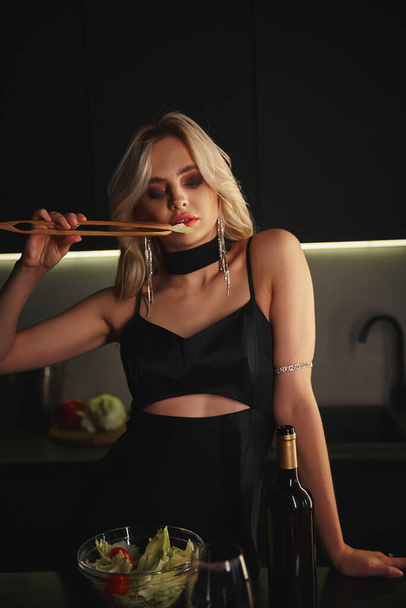 atrakcyjna młoda kobieta w czarnej sukni wieczorowej z akcesoriami za pomocą pałeczek podczas pobytu w kuchni - Zdjęcie, obraz