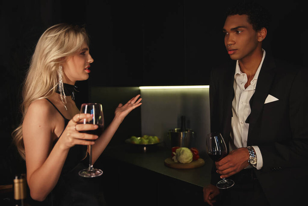 csinos multikulturális pár elegáns ruhákban vitatkoznak, és a vörösbort szemüvegben tartják. - Fotó, kép