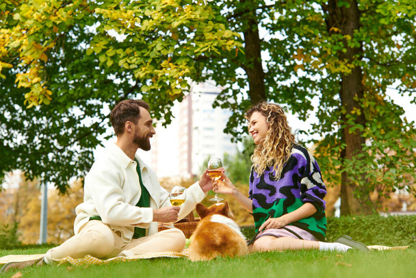 feliz pareja tintineo vasos de vino mientras está sentado en la hierba y tener picnic con perro corgi - Foto, imagen