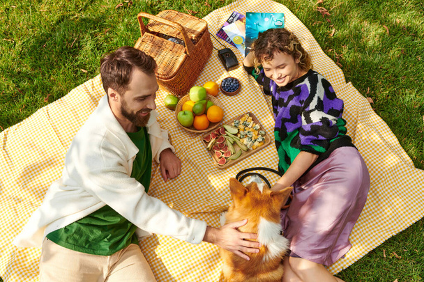 gelukkig paar genieten van picknick, rusten en knuffelen schattig corgi hond op deken naast heerlijk eten - Foto, afbeelding