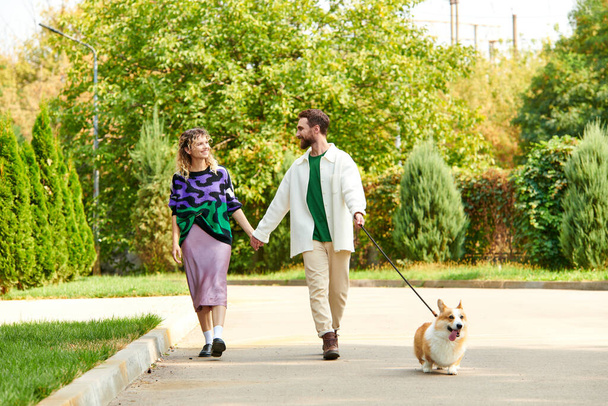 happy couple in stylish attire holding hands and walking with cute corgi dog around green trees - Photo, Image