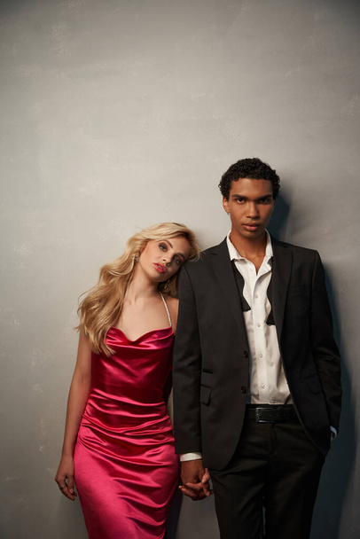 appealing young multicultural couple in elegant evening attires posing together on gray backdrop - Foto, imagen