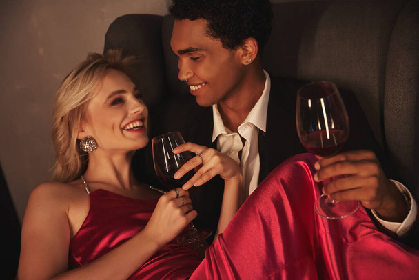 χαρούμενο ελκυστικό πολυπολιτισμικό ζευγάρι με βραδινές ενδυμασίες ποζάρουν μαζί με κόκκινο κρασί σε ποτήρια - Φωτογραφία, εικόνα