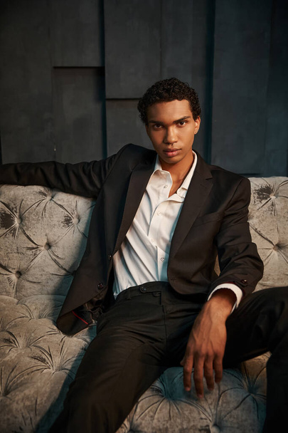 jóképű nyugodt afro-amerikai férfi modell elegáns öltönyben ül a kanapén és a kamerába néz - Fotó, kép