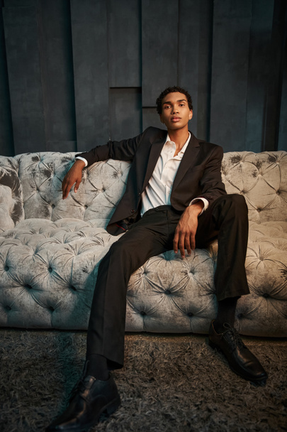 pohledný mladý africký Američan v elegantním obleku sedí na pohodlné pohovce a dívá se na kameru - Fotografie, Obrázek
