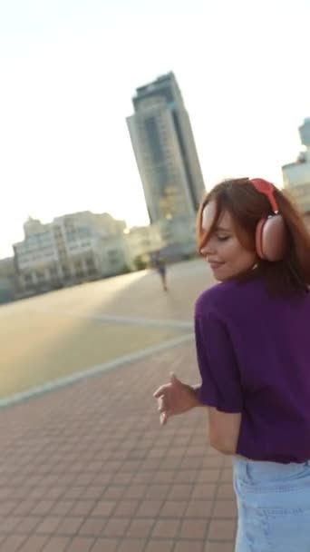 Une belle fille aux cheveux roux avec écouteurs danse sur la place du centre-ville. Images 4k de haute qualité - Séquence, vidéo