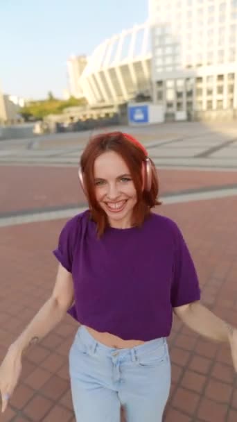 ヘッドフォンは彼女の耳を愛し,街の中央広場で魅力的な赤毛ダンス. 高品質の4k映像 - 映像、動画