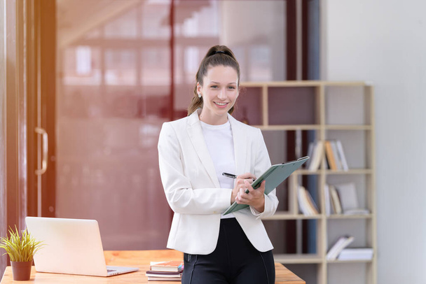 Красивая и улыбающаяся деловая женщина с бумажной работой в руках, стоящая в офисе. Случайная и успешная концепция бизнеса - Фото, изображение