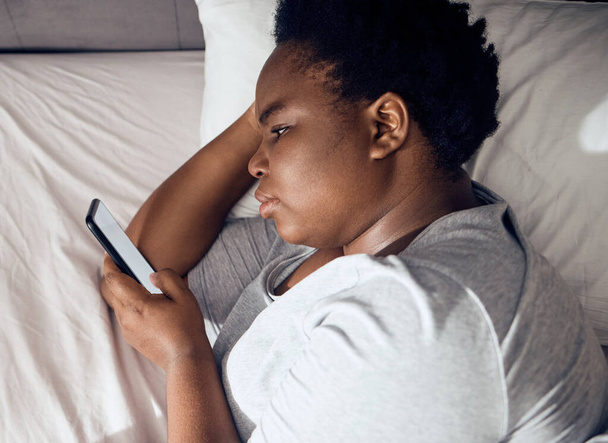 Stress, morgendliche oder schwarze Frau im Bett mit Handy für soziale Medien, die im Raum für Sucht zu Hause liegt. Schlaflosigkeit, einsame oder traurige Person, die online liest, um auf der mobilen App im Schlafzimmer mit Depressionen zu blättern. - Foto, Bild