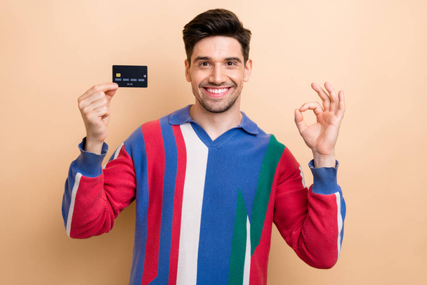 Portrait of cheerful guy beaming smile arm hold debit card demonstrate okey symbol isolated on beige color background. - Φωτογραφία, εικόνα