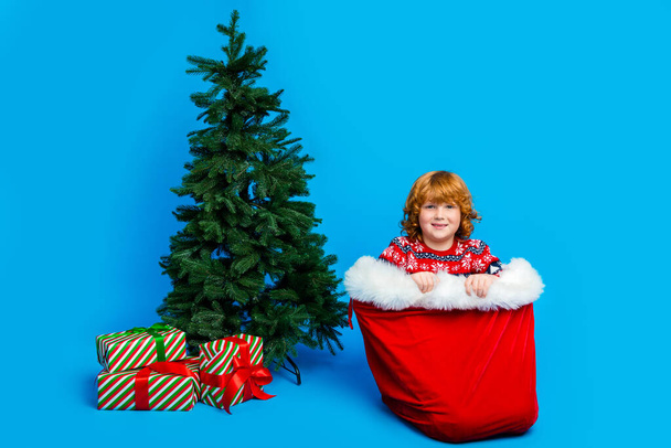 In voller Länge Foto von funky charmante kleine Junge tragen Ornament Pullover sitzt in Xmas Sack isoliert blaue Farbe Hintergrund. - Foto, Bild