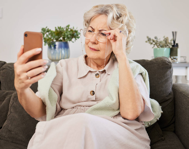 Téléphone portable, lunettes et femme âgée lisant un message texte, blog ou nouvelles en ligne sur le canapé dans le salon. Vision, technologie et sénior femme plissant tout en réseautant sur un téléphone à la maison - Photo, image