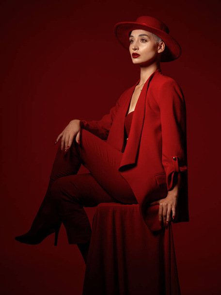 Σκέψη, μόδα και μια γυναίκα που κάθεται σε ένα κόκκινο φόντο στούντιο για κομψό ή μοντέρνο στυλ. Όραμα, τέχνη και ομορφιά σε μια καρέκλα με ένα νεαρό θηλυκό μοντέλο που αναζητούν νευρικός ή αριστοκρατικό στα ρούχα κοστούμι. - Φωτογραφία, εικόνα