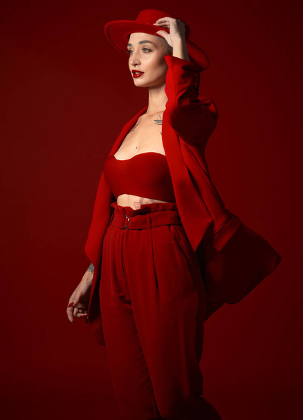 Vision, Mode und eine Frau mit Hut auf rotem Hintergrund im Atelier für eleganten oder trendigen Stil. Ästhetik, Schönheit und Zukunft mit einer selbstbewussten jungen Frau, die in einem kühnen Anzug kantig oder edel aussieht. - Foto, Bild