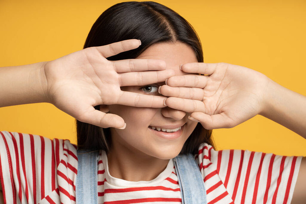 Πορτρέτο παιχνιδιάρικο χαμογελαστό έφηβος κορίτσι κρύβονται κοιτάζοντας μέσα από το χέρι ένα μάτι σε κίτρινο φόντο. Μπρουνέτ χαρούμενος έφηβος παίζει κρυφτό. Banner, αφίσα για διαφήμιση, έννοια μάρκετινγκ. - Φωτογραφία, εικόνα