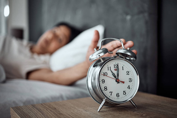 Alarm, sleeping and a hand with a clock for awake, oversleep or tired in the bedroom of a home. House, ring and a closeup of a person or woman in bed for snooze, rest or waking up lazy in a house. - Photo, Image