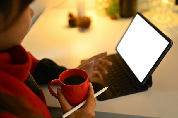 Jonge vrouw met een kopje hete koffie en met behulp van digitale tablet in ingerichte kamer met warm licht. - Foto, afbeelding