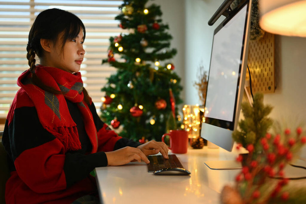 Sıcak örgü süveter giyen genç bir kadın süslü Noel ağacının yanında oturuyor ve bilgisayar kullanıyor.. - Fotoğraf, Görsel