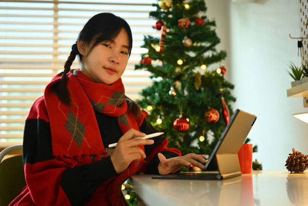 Hübsche junge Frau in warmer Kleidung mit digitalem Tablet sitzt am geschmückten Weihnachtsbaum im Wohnzimmer. - Foto, Bild