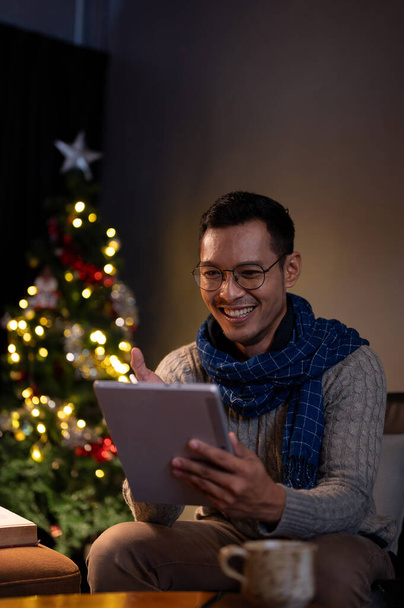 Ένα πορτραίτο ενός όμορφου, ευτυχισμένου Ασιάτη με ζεστά ρούχα χρησιμοποιώντας το ψηφιακό του τάμπλετ ενώ χαλαρώνει στο σαλόνι του τη νύχτα των Χριστουγέννων. - Φωτογραφία, εικόνα