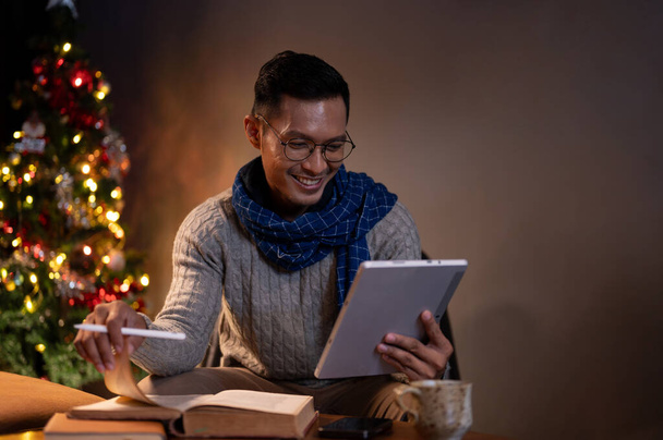 Egy boldog ázsiai férfi kényelmes pulóverben és sálban használja a digitális tabletjét, és könyvet olvas egy sötét nappaliban karácsony este, egyedül ünnepli a karácsony estét.. - Fotó, kép