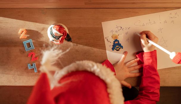 criança em um chapéu vermelho escreve uma carta ao Papai Noel sonhadoramente pensando em presentes. Os números para 2024 estão próximos. Lista de desejos de presentes. À espera de um milagre de Natal. Vista de cima. Feriados de Ano Novo - Foto, Imagem