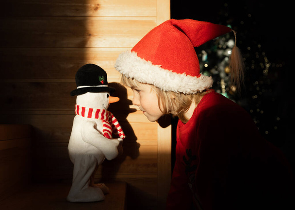 Noel Baba şapkası takmış neşeli çocuk oyuncak bir kardan adamla burun buruna çarpışırken eğleniyor. Noel arifesinde, tatil beklentisiyle evde oynuyoruz. Güneş ışığıyla nesneleri renklendiriyor - Fotoğraf, Görsel
