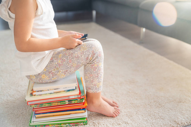 Petite fille s'assoit sur une pile de livres de contes de fées pour enfants et regarde des dessins animés sur smartphone aux rayons du soleil. Photo de haute qualité - Photo, image
