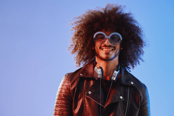 Portrait of a stylish man with curly hair with glasses and headphones on a blue background multinational, colored light, black leather jacket trend, modern concept. High quality photo - Fotografie, Obrázek