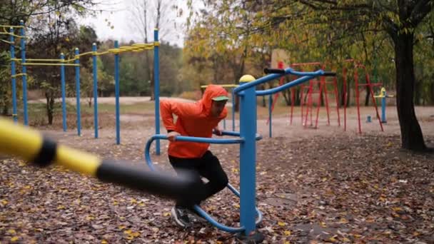 young man exercising outdoor at sports ground, pumps up muscles using equipment in cold autumn day. - Footage, Video