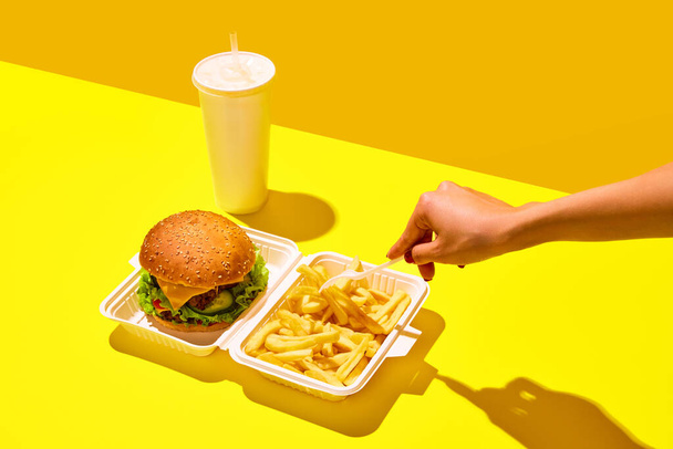 Распаковываю еду на дегустацию. Неузнаваемая женщина ест гамбургер с содовой и жареным картофелем в бумажных коробках на ярком желтом фоне. Концепция нездоровой пищи, меню, доставка, питание, забрать. Объявление - Фото, изображение