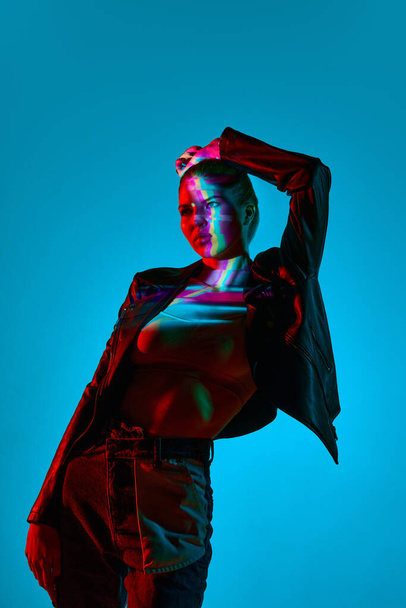 Attractive young woman in stylish casual clothes standing against blue background with multicolored neon lights reflection. Concept of modern art, beauty, style, futurism and cyberpunk, creativity - Photo, image