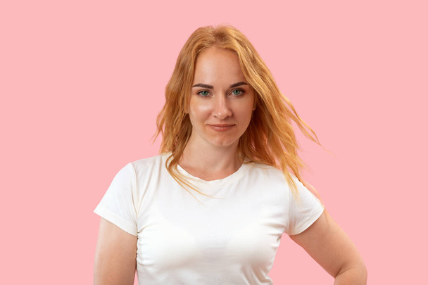Beyaz tişörtlü, pembe arka planlı, uzun dalgalı saçlı, çekici, kızıl saçlı bir kadının portresi. Güzel kendine güvenen kadın. - Fotoğraf, Görsel