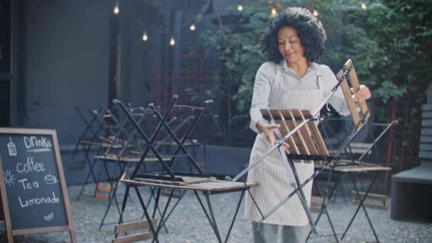 Beautiful African American woman arranging chairs down on floor before starting of working day in cafe. Preparing terrace for serving upcoming customers. Enjoying working in morning. Workday concept. - Footage, Video