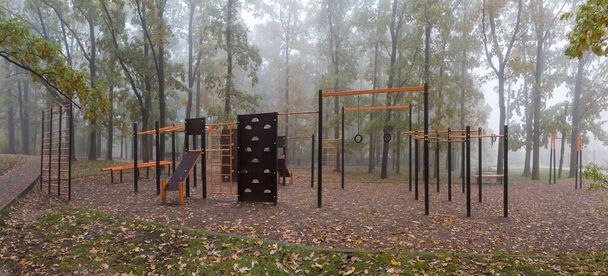 秋の霧の公園,パノラマビューの木の間の自由な運動トレーニングそしてフィットネスのための装置が付いている空の公共の屋外スポーツ区域 - 写真・画像