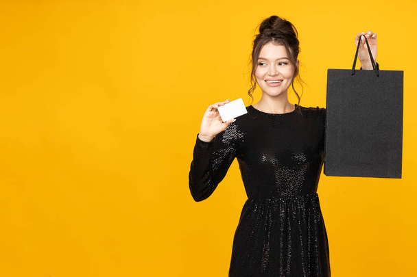 Happy young woman with card and paper bag in hands on yellow background, space for text - Photo, Image