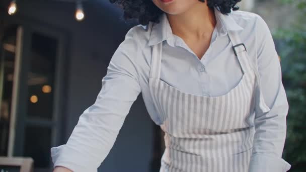 Czarująca Afroamerykanka baristka w niebieskiej koszuli i fartuchu pracująca sama rano na tarasie kawiarni. Stoły czyszczące z tkaniny z bakterii. Przygotowywanie się do przyjmowania klientów. - Materiał filmowy, wideo