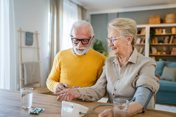 Senioren Paar Frau messen Blutdruck, während ihr Mann alten kaukasischen Mann sitzen neben am Tisch zu Hause echte Menschen Familie Senioren Ruhestand und Gesundheitskonzept - Foto, Bild