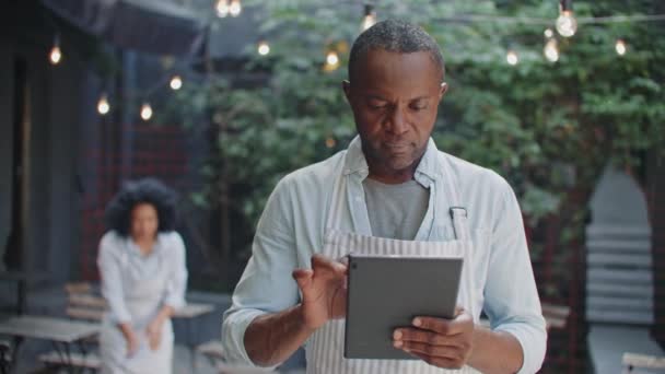 Komea Afrikkalainen amerikkalainen mies omistaja pieni kahvila käyttäen tabletin laitteen seisoessaan terassilla. Kiireinen miespuolinen laskenta tai ruoan tilaaminen digitaalisella laitteella, kun taas nuoret tarjoilijat puhdistavat pöytää taustalla - Materiaali, video