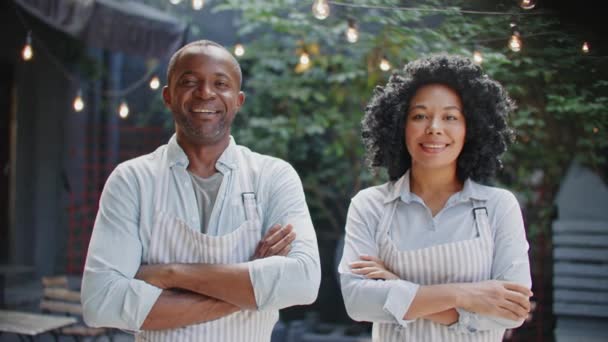 Zwei afroamerikanische Arbeiter mit Schürzen, die sich vor der Kamera die Hände kreuzen. Positive Menschen, die bei der Arbeit im modernen Café fröhlich lächeln. Partnerschaft. Coworker-Konzept. - Filmmaterial, Video