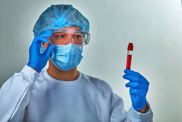 Γιατρός σε μάσκα κοιτάζοντας το σωλήνα εξέταση αίματος για coronavirus COVID19, HIV, Έμπολα ή άλλη επικίνδυνη λοίμωξη. Ιατρικό υπόβαθρο, όργανα, πρότυπο, ταπετσαρία. Έννοια της νόσου του Coronavirus - Φωτογραφία, εικόνα