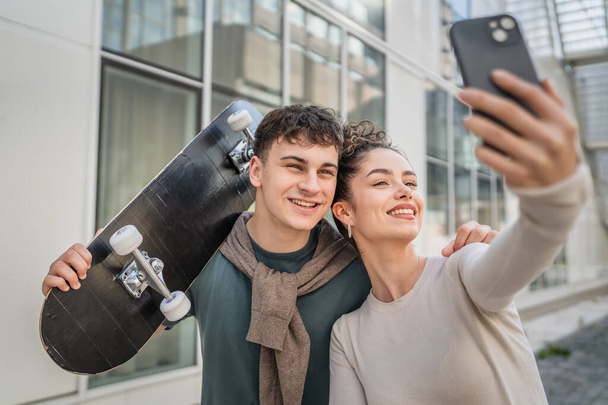 młody mężczyzna i kobieta nastolatek para kaukaski brat i siostra zrobić selfie autoportret przez nowoczesny budynek w dzień szczęśliwy uśmiech nosić deskorolka prawdziwe ludzie rodzina miłość koncepcja przestrzeń - Zdjęcie, obraz