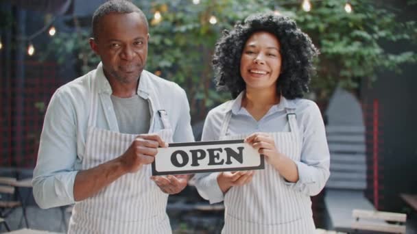 Satisfied African American people holding open sign while standing on cafe terrace. Joyfully smiling at camera. Happy about working together at cozy bar. Getting ready to hard working day. - Footage, Video
