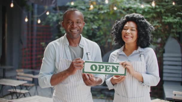 Zwei afroamerikanische Kellner mit Schürzen halten ein offenes Schild auf der Caféterrasse. Glückliche Arbeiter lächeln vor Freude, während sie vor der Theke stehen. Bereiten Sie sich darauf vor, Kunden zu empfangen. Konzept des Arbeitstages. - Filmmaterial, Video