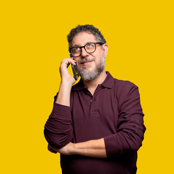 Ein nachdenklicher Mann mittleren Alters mit Brille und grauem Bart führt ein Telefongespräch, trägt ein kastanienbraunes Hemd vor leuchtend gelbem Hintergrund und strahlt Neugier und Konzentration aus.. - Foto, Bild