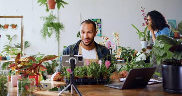 Handsome man gardener in apron using phone and recording blog video with clients. Young adult content creator influencer blogger recording a video talking about indoor plants care from workplace. - Foto, Imagem