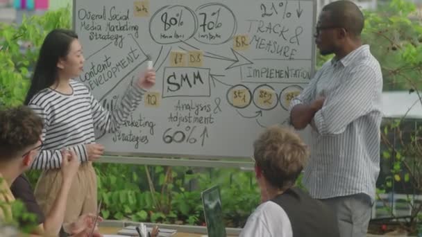Hombre negro y mujer asiática señalando la presentación en pizarra blanca y hablando con colegas durante la reunión de negocios al aire libre - Metraje, vídeo