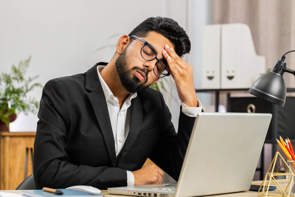 Уставший индийский бизнесмен, страдающий от головной боли, напряженности и мигрени, стресса на рабочем месте домашнего офиса. Бородатый брокер-фрилансер работает над ноутбуком. Переработка - Фото, изображение