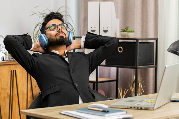 Happy smiling Indian businessman listening music on headphones taking break relaxing leaning on chair after working at office workplace. Male Hindu freelancer in formal suit puts hands behind head - Photo, Image