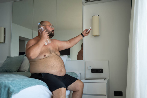 コールドウォータースイーツのボトルを持つ裸の肥満男性は,ホテルの部屋に座っている暖かさの異常な程度に対処しようとリモートコントロールでエアコンを調整します. 脂肪の男性は熱から死にます. - 写真・画像